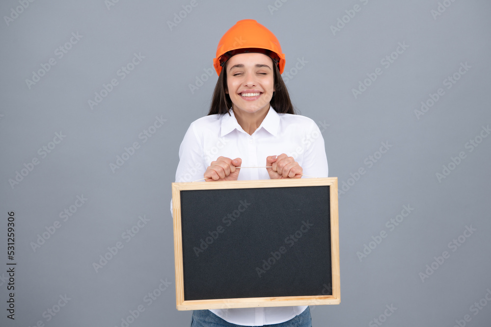 Young beautiful smiling architect woman in hard hat helmet hold board over gray background. Funny emoticons girl in a construction helmet. Young beautiful smiling architect woman in hard hat helmet hold board over gray background. Funny emoticons girl in a construction helmet.