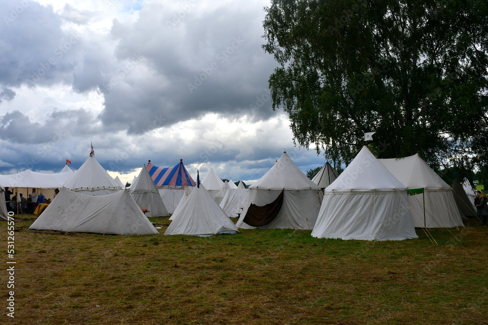 A close up on a tent made out white cloth with a wooden frame and ropes supporting it seen during a big medieval folklore festival or fair right before a massive thunderstorm and rainfall