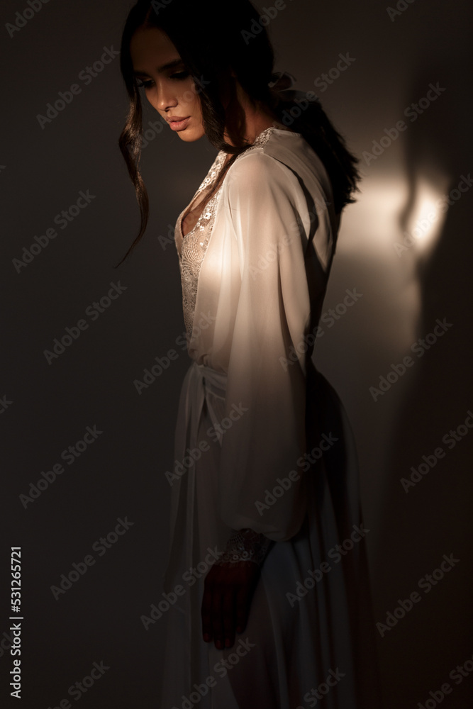 Attractive bride wearing white transparent robe decorated with guipure in the dark studio. Fashion bride clothes.
