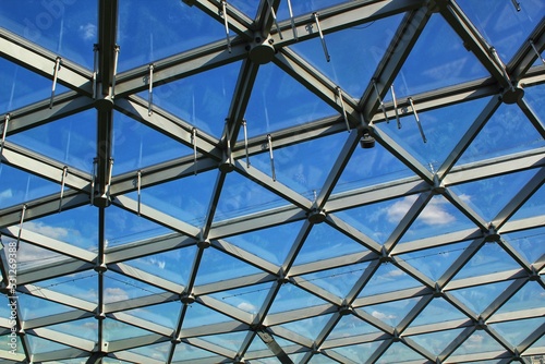Transparent glass dome roof of modern design style pavilion building with geometric metal construction pattern. View of blue sky and clouds. Abstract structure triangle high-tech architecture window.