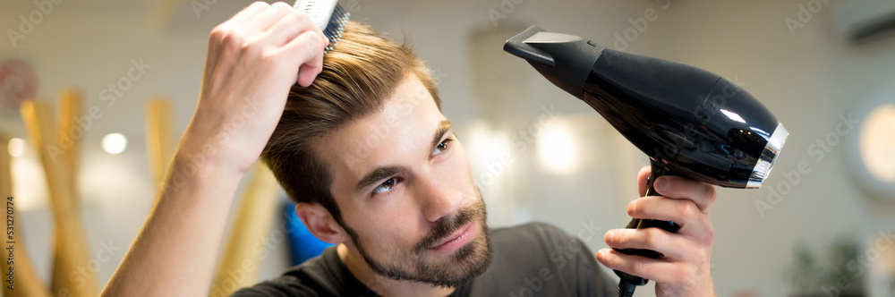 Fototapeta premium Young man hairdresser drying and combing his own hair in his hair salon