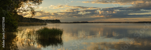 a large lake with coastal reeds, trees and the reflection of the sky in the water in the warm light of the morning dawn. widescreen panoramic view