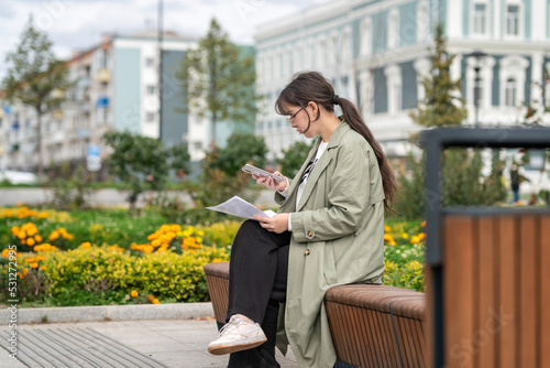 A girl sits on a bench on the street with documents in her hands and using her phone