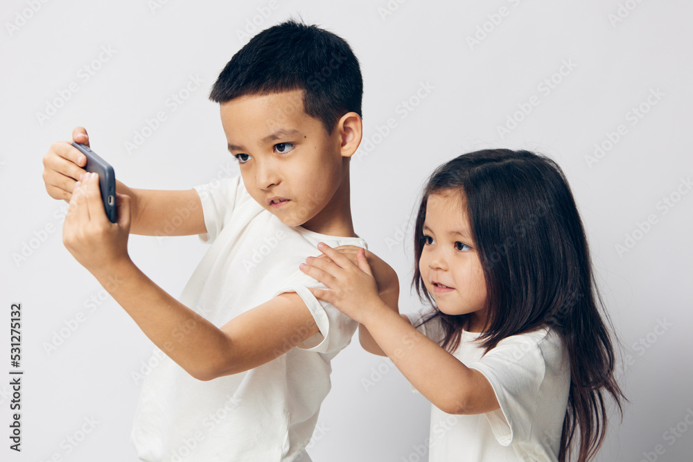 brother and sister take a selfie on their fashionable smartphone while standing in white T-shirts on a light background sideways to the camera brother and sister take a selfie on their fashionable smartphone while standing in white T-shirts on a light background sideways to the camera
