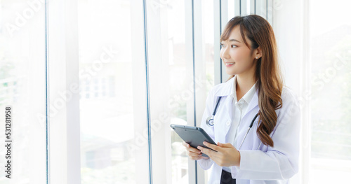Beautiful and cheerful good mood young lady asian doctor dressed uniform smiling arm stethoscope while using tablet on lobby hospital background. © aekkorn