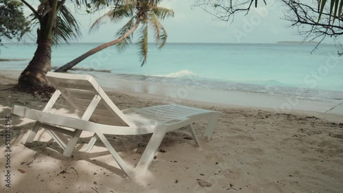 View from behind with a selective focus on a daybed on the sand of a luxury resort opposite teal ocean water with slow-motion waves and splashes in the defocused background; luxury resort, Maldives