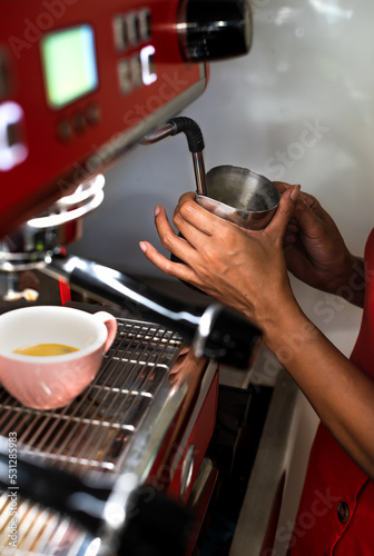 hands of a barista holding the metal jug where she is heating the milk for her customer s coffee. the machine and her uniform are red. there is a colorful cup with coffee. vertical photograph.