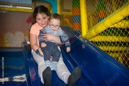 In the playroom on the slide, the sister holds her little brother in her arms and slides down.