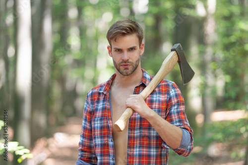 Unshaven guy in lumberjack shirt holding axe on shoulder forest background