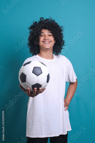 Soccer fan with curly hair wig supporting favorite team. Portrait of happy funny excited cheerful man holding football, looking at camera and smiling © Gatot