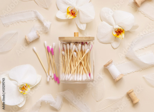 Bamboo cotton buds in a jar near white orchid flowers on light beige. Eco friendly skincare product