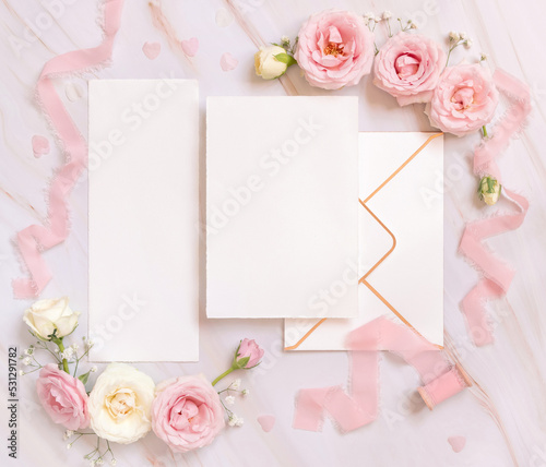 Blank paper cards between pink roses and pink silk ribbons on pink top view, wedding mockup