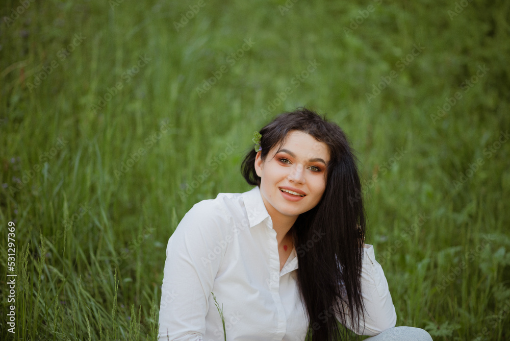 Sexy brunette sits on the grass in a summer park in a white blouse and jeans Sexy brunette sits on the grass in a summer park in a white blouse and jeans