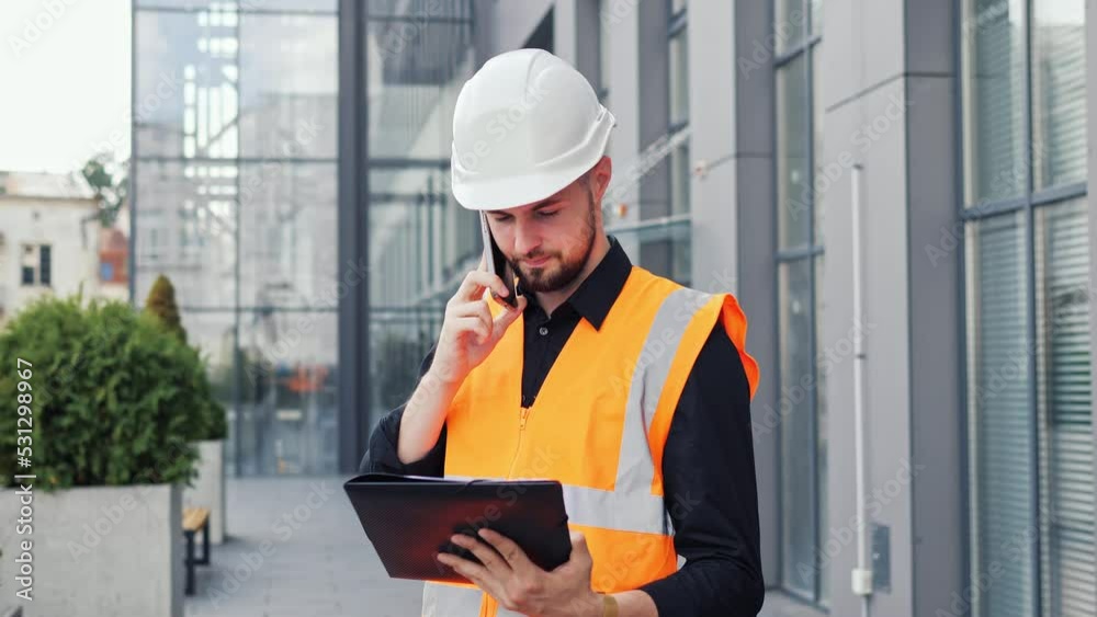Young caucasian man architect designer holding document project talking with a contractor by mobile phone. Businessman discusses details of project with customer or boss, business conversation. Young caucasian man architect designer holding document project talking with a contractor by mobile phone. Businessman discusses details of project with customer or boss, business conversation.