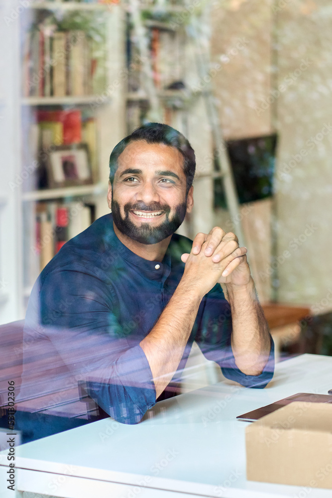 Smiling handsome rich bearded indian ethnic business man, successful wealthy eastern professional businessman looking away laughing sitting at work desk. Vertical authentic shot through glass Smiling handsome rich bearded indian ethnic business man, successful wealthy eastern professional businessman looking away laughing sitting at work desk. Vertical authentic shot through glass