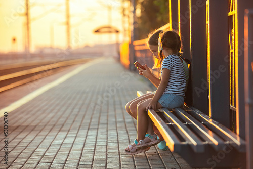 two little girls tourist sisters are sitting on the bench of the railway station and watching the phone while waiting for the electric train in the evening in the rays of the setting sun
