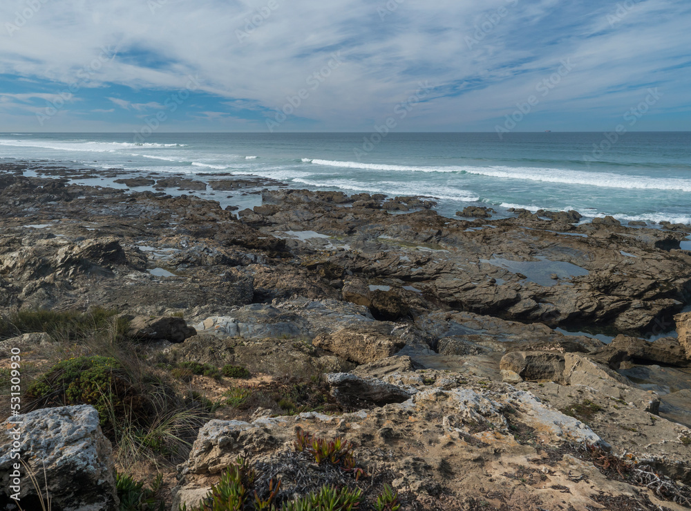 View sea shore with ocean waves, sharp rocks, stones and green vegetation at Rota Vicentina wild coast near Porto Covo, Portugal. View sea shore with ocean waves, sharp rocks, stones and green vegetation at Rota Vicentina wild coast near Porto Covo, Portugal.