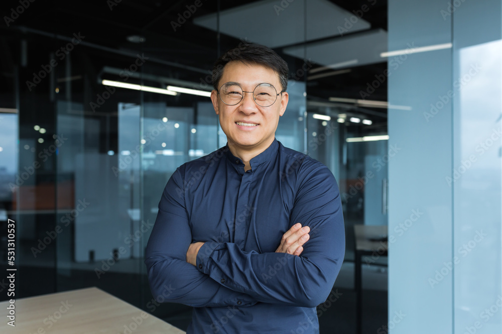Portrait of happy and successful asian businessman man in glasses smiling and looking at camera, manager business owner with crossed arms near window working inside modern office Portrait of happy and successful asian businessman man in glasses smiling and looking at camera, manager business owner with crossed arms near window working inside modern office