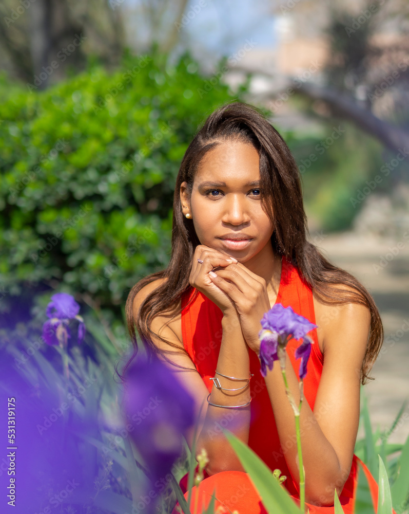 Fototapeta premium Joven adolescente de Colombia, mirando a cámara sentada entre las flores lilas del campo, con árboles verdes desenfocados al fondo en un retrato de primer plano, en la primavera de 2021