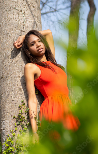 Joven colombiana delgada, mulata con vestido rojo y pelo largo oscuro, apoyada en un tronco con la mano arriba y hojas verdes en la primavera de 2021. photo