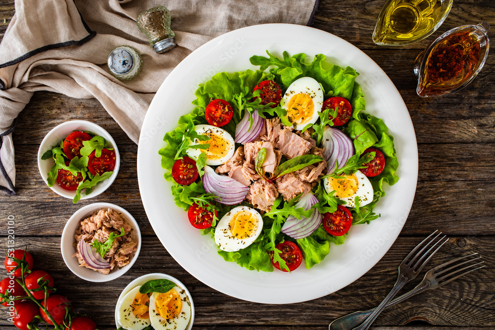 Naklejka premium Tuna salad - tuna, hard boiled eggs, cherry tomatoes, lettuce and onion on wooden table 