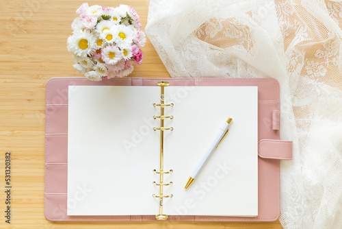 Flat lay, top view of a pastel pink diary, flower bouquet and stationery on a wooden desk.