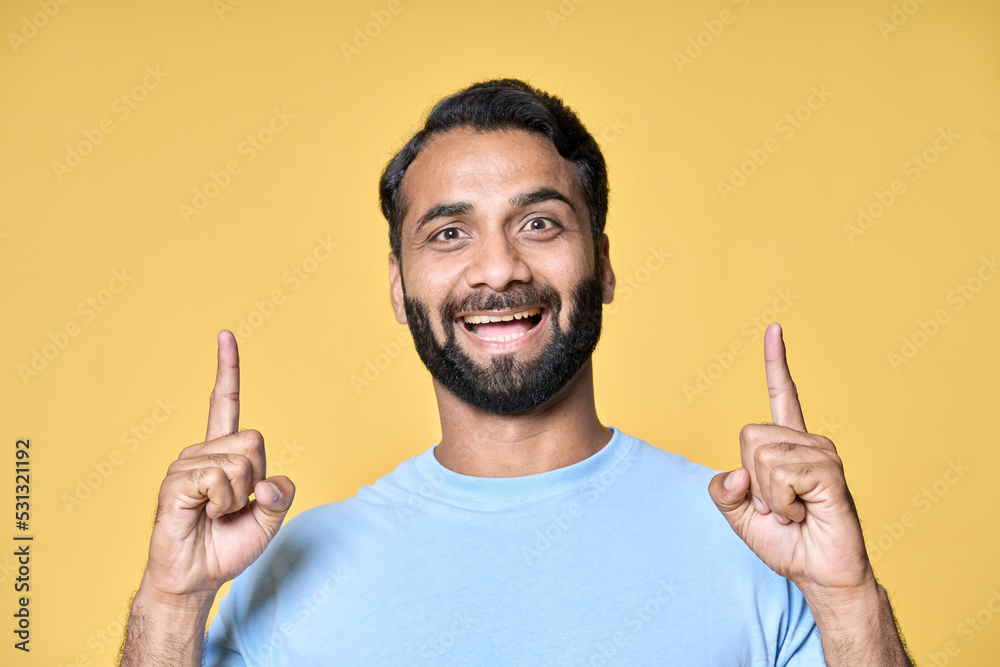Excited indian man looking at camera pointing up with fingers hand gesture indicating advertising new product ecommerce promo, presenting sale offer standing isolated on yellow background. Excited indian man looking at camera pointing up with fingers hand gesture indicating advertising new product ecommerce promo, presenting sale offer standing isolated on yellow background.