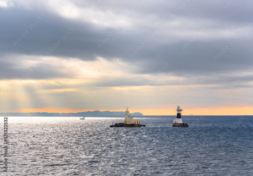 Lighthouse and oceanographic observation station of Ashikashima in the sea of the Uraga channel of Tokyo bay with a boat sailing along the Boso peninsula coasts in the morning light below a cloudy sky