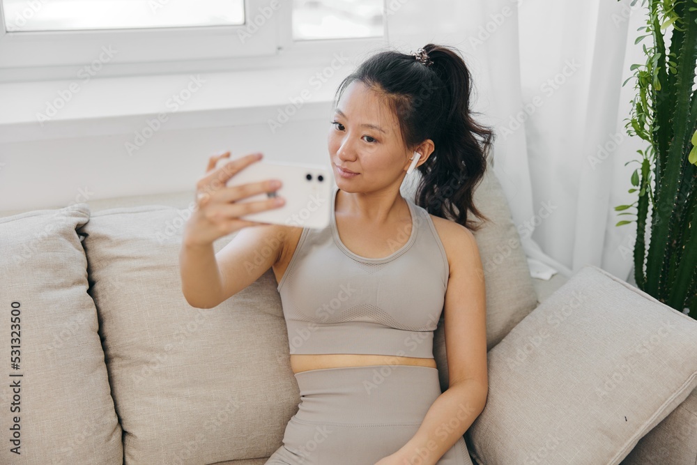 Athletic Asian woman in yoga costume blogger films herself on her phone camera sitting on the couch and smiling with headphones, freelance work from home, recording sports workout lifestyle body care Athletic Asian woman in yoga costume blogger films herself on her phone camera sitting on the couch and smiling with headphones, freelance work from home, recording sports workout lifestyle body care
