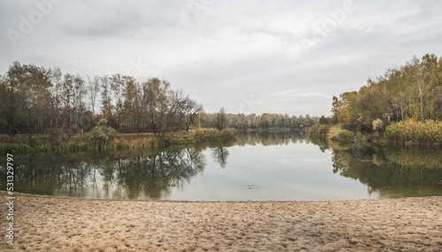 The bay of the steppe river in autumn cloudy weather along the banks of which trees with yellowed October foliage grow, in the foreground there is a beach