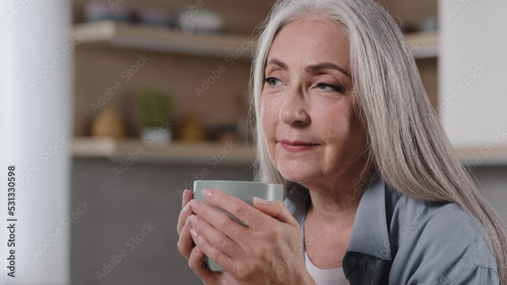Dreamy mature woman holding cup of hot beverage warming hands on mug preventing flu looking away drinking morning coffee positive dreaming pleasant memories happy elderly grandmother relaxing at home Dreamy mature woman holding cup of hot beverage warming hands on mug preventing flu looking away drinking morning coffee positive dreaming pleasant memories happy elderly grandmother relaxing at home