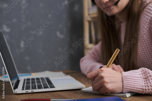 A girl with a headset works at a laptop, noting something with a pen in a notebook. The topic of online work, support, training or remote communication. Close up