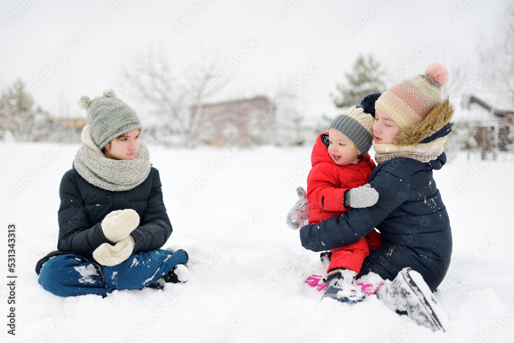 Two cute young sisters and their toddler brother having fun in snow covered park on chilly winter day. Children exploring nature.