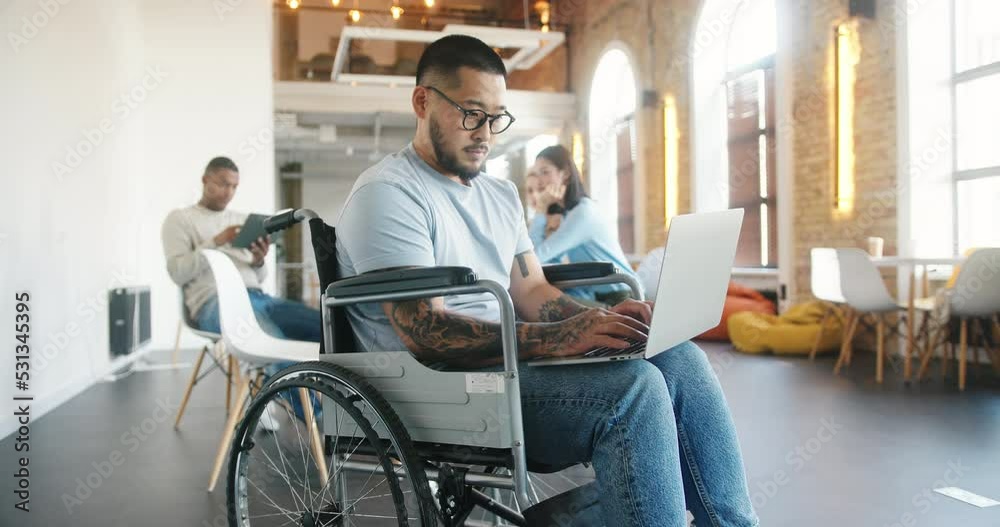 Latin man in wheelchair works on laptop against colleagues Latin man in wheelchair works on laptop against colleagues