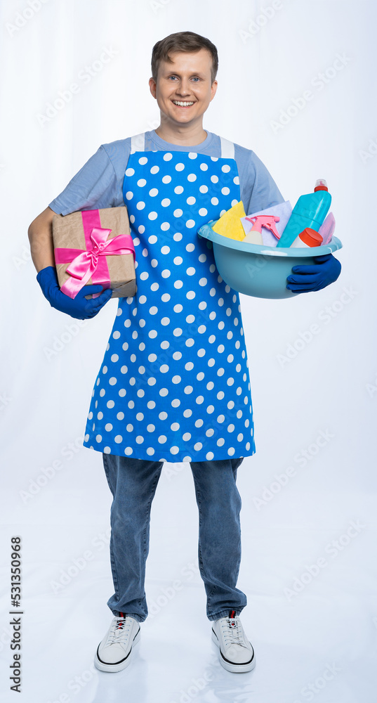 a man in a blue apron, rubber gloves, an orange t-shirt, jeans and white sneakers holds a basin with detergents and a gift for a birthday or women's day. isolated white background