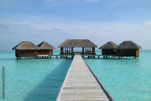 Tropical resort in Maldives showing overwater huts and bungalows with a long pier, thatched-roofs, crystal clear water and blue sky