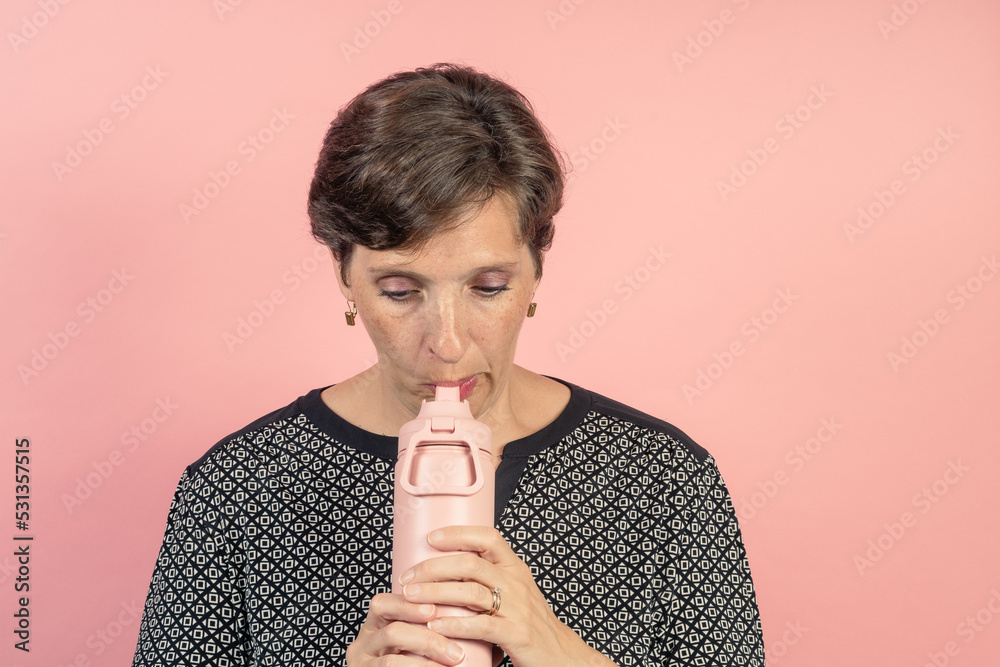 Brunette woman in her 50s in the studio with a pink background about to drink from a flask Brunette woman in her 50s in the studio with a pink background about to drink from a flask