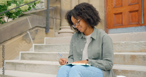 African American young woman with curly hair and glasses, seats on the stairs outdoors and writing in her notepad with a joy smile