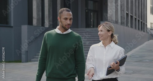 Multiracial Positive Man and Adult Woman Holding Laptop and Talking. Businesspeople Outside at the Street Discussing Business Plans with Each Other while Walking