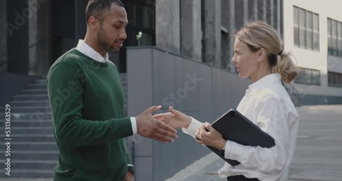 Multiracial Young Man and Senior Woman Discussing Project. Elegant Colleagues Standing on Street with Laptop. Urban Background 