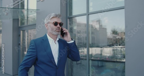 Senior Businessman Talking Mobile Walking On Street. Business Person Holding Smartphone On Go During Break. Mature Guy Call To Colleague Near Office Building. Business and Lifestyle Concept 