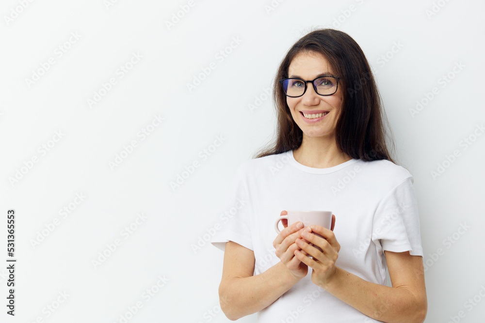 a sweet, beautiful, pleasant woman in glasses and a white t-shirt stands holding a cup in her hands and smiles sweetly.Horizontal photo on a light background a sweet, beautiful, pleasant woman in glasses and a white t-shirt stands holding a cup in her hands and smiles sweetly.Horizontal photo on a light background