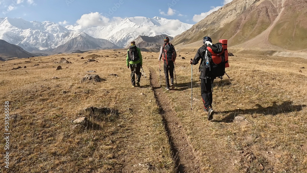 Three climbers with backpacks walk along the path past the majestic Pamir Mountains. View from the back. Beautiful summer mountain landscape.