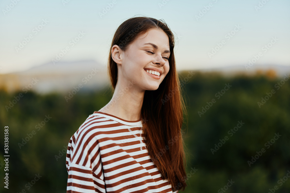 Close-up portrait of a young woman with a beautiful smile with teeth in a striped t-shirt against the background of trees
