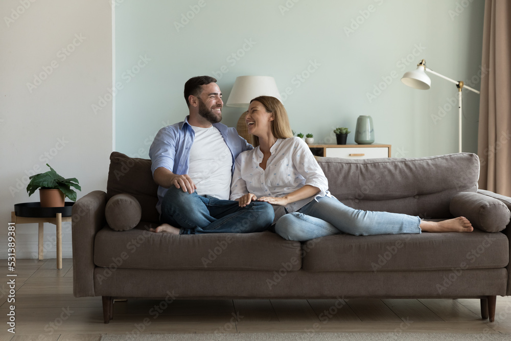 Happy joyful married couple resting on comfortable couch together, enjoying being at new cozy home, talking, laughing, having fun. Relationship, love, real estate for family concept - obrazy, fototapety, plakaty 
