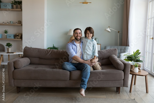 Happy handsome dad and little son kid sitting on sofa at home, hugging, looking at camera, enjoying friendship, family leisure together. Joyful father holding boy in arms. Wide shot portrait © fizkes
