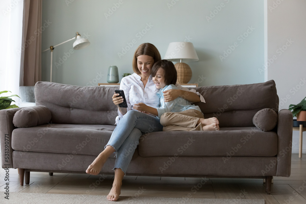 Millennial mom hugging little son on couch, using online app on mobile phone, talking on video call, shopping on Internet, showing smartphone to happy boy. Family, communication concept. Full length Millennial mom hugging little son on couch, using online app on mobile phone, talking on video call, shopping on Internet, showing smartphone to happy boy. Family, communication concept. Full length