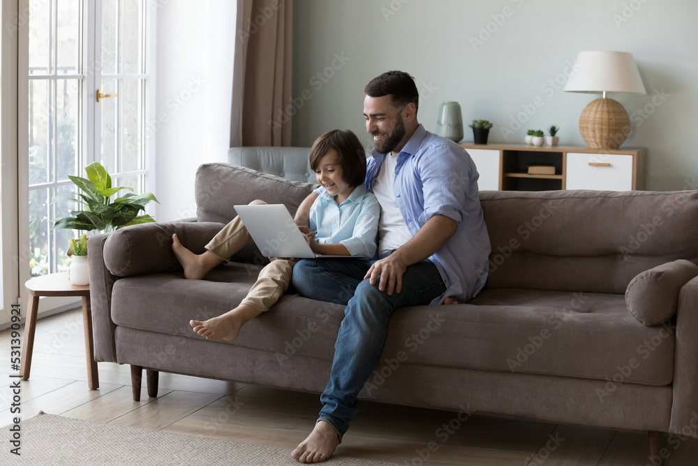 Happy millennial father hugging curious gen Z son kid, sitting on couch with laptop. Cheerful dad and little gamer boy playing virtual video game on computer. Fatherhood, family communication concept Happy millennial father hugging curious gen Z son kid, sitting on couch with laptop. Cheerful dad and little gamer boy playing virtual video game on computer. Fatherhood, family communication concept