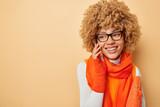 Horizontal shot of good looking woman smiles happily touches face gently looks gladfully away wears transparent eyeglasses warm orange scraf around has happy mood isolated over beige background