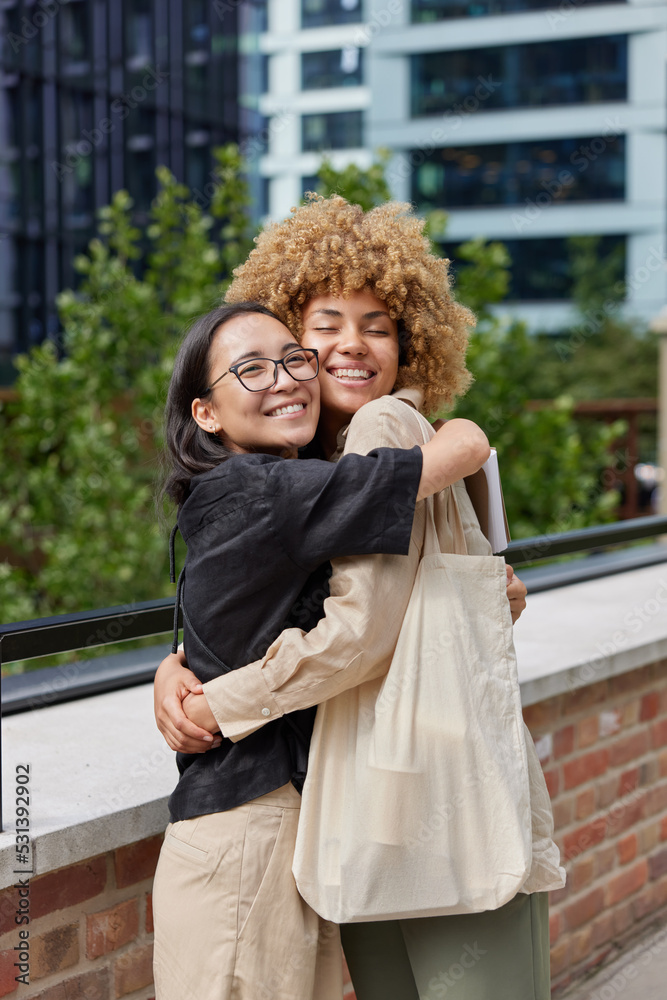 Fototapeta premium Vertical shot of happy diverse women glad to meet each other embrace and smile joyfully dressed in stylish clothes carry bag pose outdoors against blurred background. Best friends and relationship